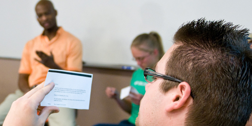 Close up of a male student reading a card, while in the back there is a male speaker talking.