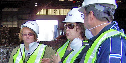 Three people with hard hats, surgical masks and safety vests on in trash facility