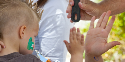 Pictured is a small boy with a dinosaur painted on his cheek. He is looking in the mirror and high five-ing someone.