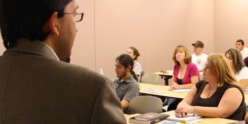 Person standing in front of a class presenting while the audience looks at the board/screen projector.