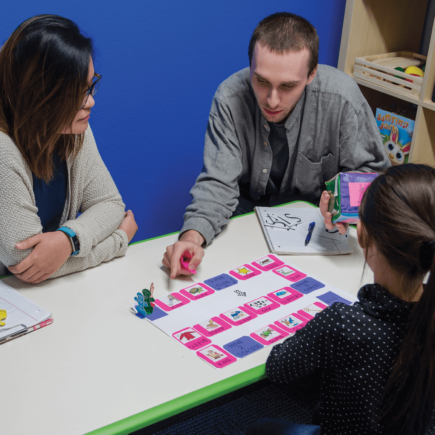 Adult female student watches as Adult male instructor teaches Female child how to play a board game