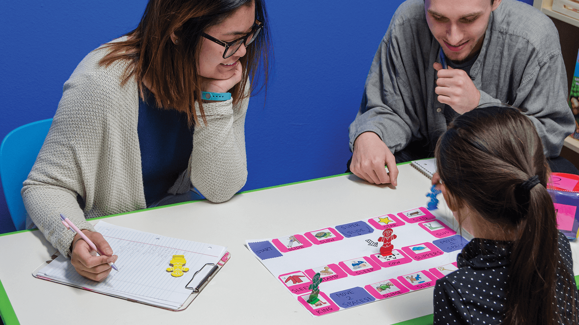 Adult male playing a board game with female child, as adult female students watches and takes notes