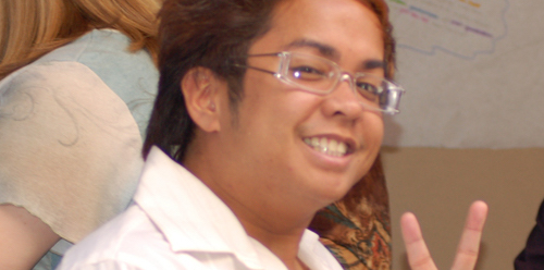 Picture of student with glasses smiling at camera holding up peace sign with fingers