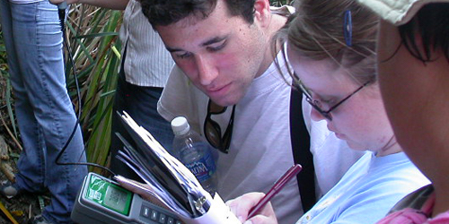 Students looking at what female student is writing in her notebook in the field