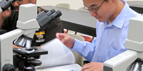 Male student reading book on desk in front of microscope