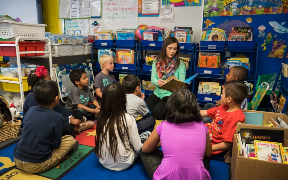 classroom with students sitting on the floor listening to adult read story